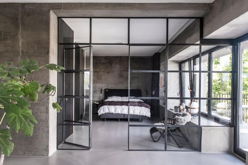 Sypialnia w stylu industrialnym z betonowymi ścianami i podłogą, oddzielona od salonu przeszkloną ścianą z czarnymi szprosami. W tle widoczne łóżko z białą pościelą i czarnym zagłówkiem, a po...