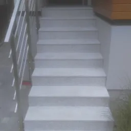 Zewnętrzne schody wejściowe wyłożone jasnoszarym kamieniem z metalową balustradą, prowadzące do ciemnych drzwi wejściowych.