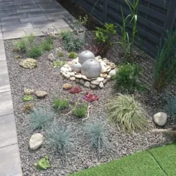 Ogród z dekoracyjnym oczkiem wodnym z kamiennych kul, otoczonym drobnymi kamieniami i roślinami, obok kostki brukowej i sztucznej trawy, na tle ciemnego drewnianego płotu.