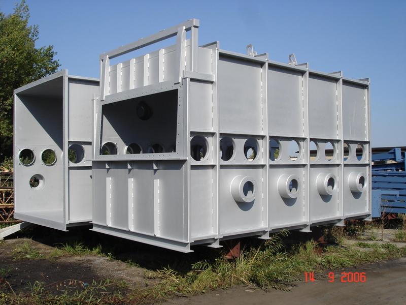 Szara, spawana konstrukcja stalowa z otworami, prawdopodobnie fragment większej instalacji przemysłowej, na zewnątrz w słoneczny dzień.