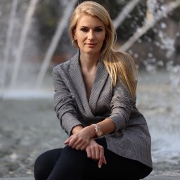 Katarzyna Szczęsna Ubezpieczenia - Kobieta w szarym żakiecie siedzi na tle fontanny, prezentując ofertę ubezpieczeniową.