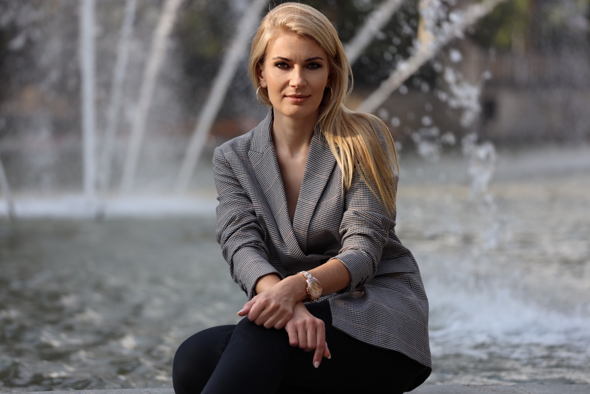 Kobieta w szarym żakiecie siedzi na tle fontanny, prezentując ofertę ubezpieczeniową.