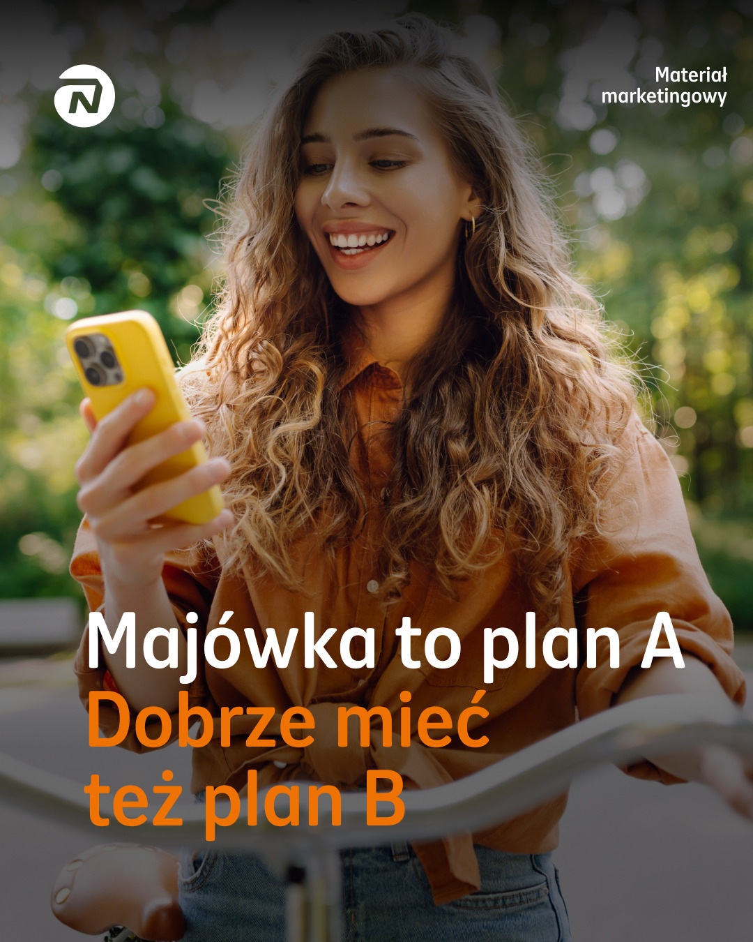 Uśmiechnięta kobieta z telefonem na rowerze, tekst: Majówka to plan A, dobrze mieć też plan B. Materiał marketingowy.