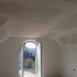 Pokój w trakcie prac wykończeniowych, sufit i ściany po szpachlowaniu, zabezpieczona folią podłoga, widok na zewnątrz przez otwarte, łukowe drzwi balkonowe.