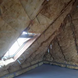 Poddasze z oknem dachowym w trakcie ocieplania wełną mineralną, widoczne metalowe uchwyty mocujące izolację do drewnianej konstrukcji dachu.