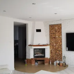 Jasne wnętrze salonu z kominkiem obłożonym białym kamieniem i dekoracją z drewna za telewizorem na ścianie z białej cegły.