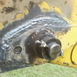 Spoina na żółtym elemencie konstrukcyjnym maszyny budowlanej, widoczny sworzeń z nakrętką, ślady smaru i zabrudzeń.