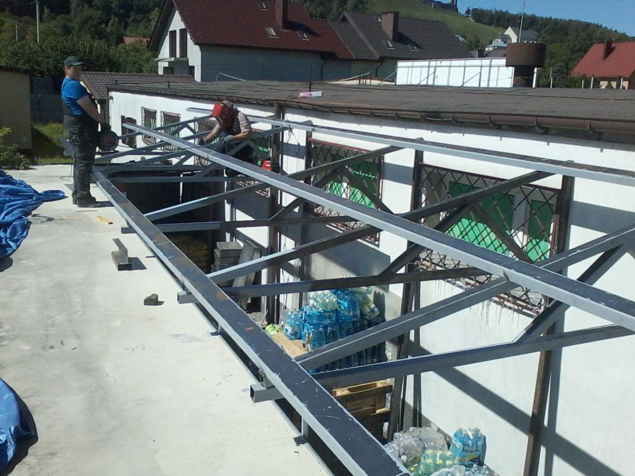 Dwóch pracowników montuje metalową konstrukcję szkieletową na dachu budynku, widoczne narzędzia pracy i zapas wody w butelkach.