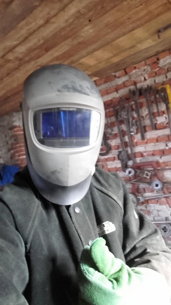 Spawacz w masce ochronnej i roboczej kurtce, w tle ceglana ściana z narzędziami.