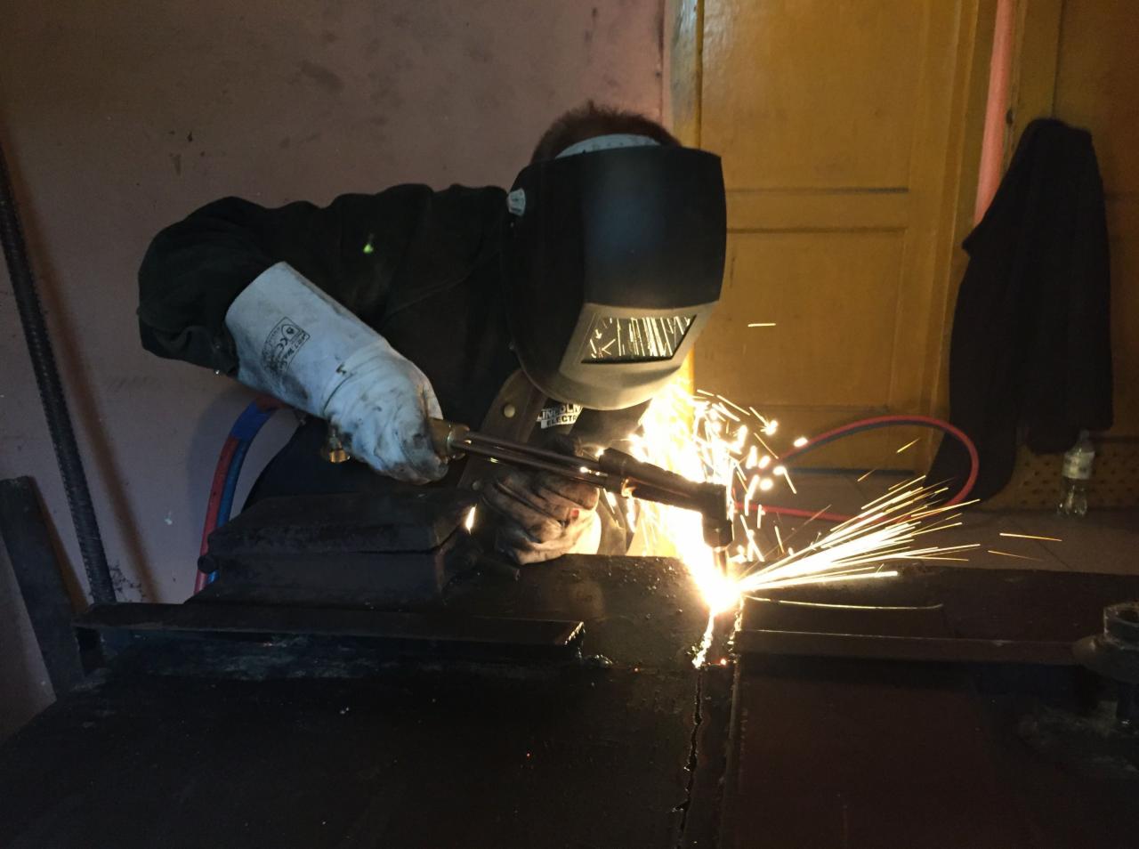 Pracownik w masce spawalniczej łączy metalowe elementy za pomocą palnika, iskry rozbryzgują się w jasnym świetle.