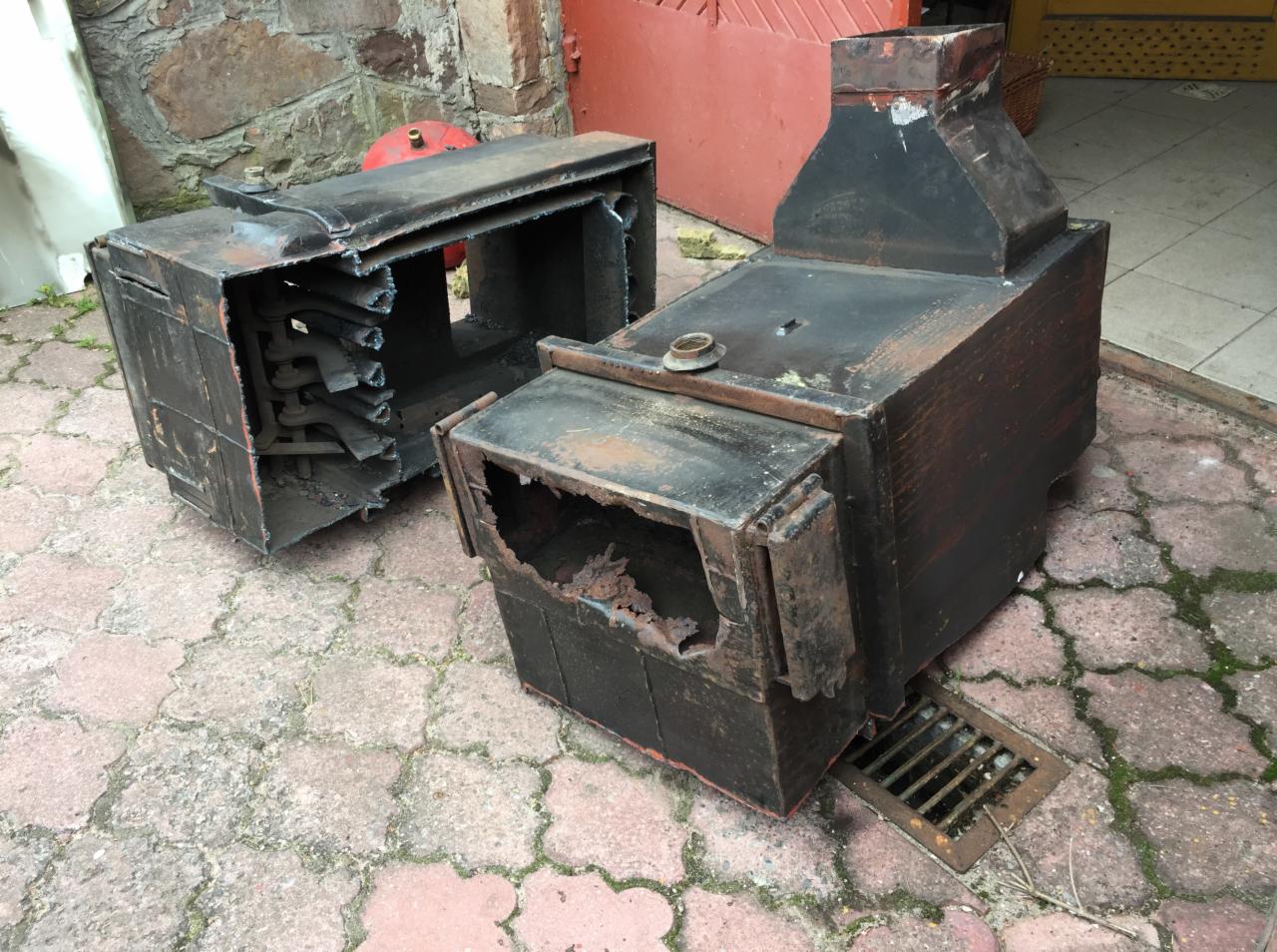 Zdemontowany, skorodowany piec centralnego ogrzewania, podzielony na dwie części, leżący na brukowanym podwórku, widoczne uszkodzenia i rdzawy nalot.