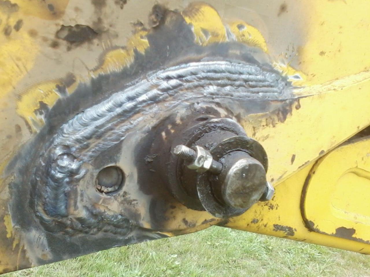 Spoina na żółtym elemencie konstrukcyjnym maszyny budowlanej, widoczny sworzeń z nakrętką, ślady smaru i zabrudzeń.