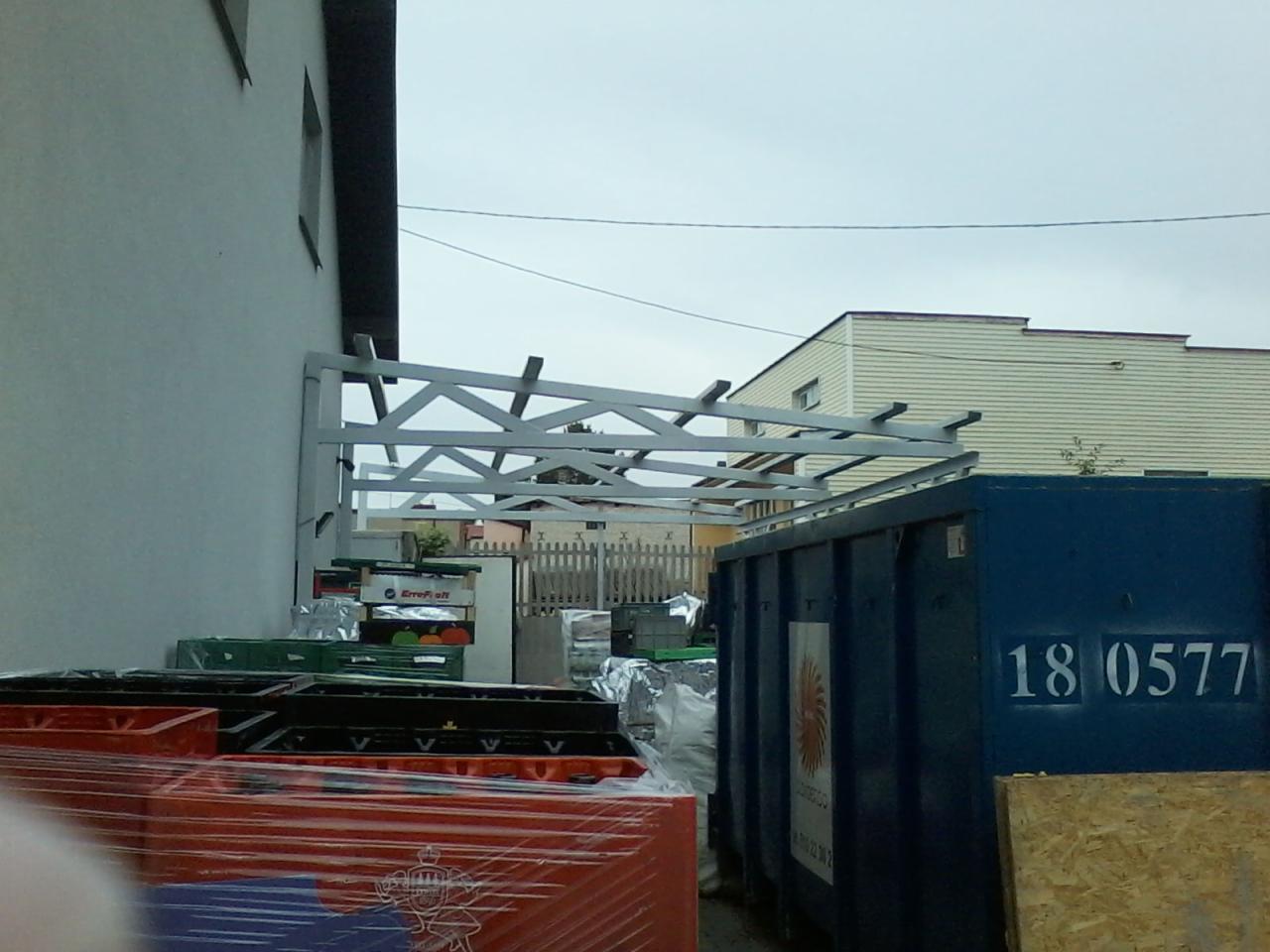 Stalowa konstrukcja zadaszenia przy budynku, widoczny kontener na śmieci i skrzynki w folii.
