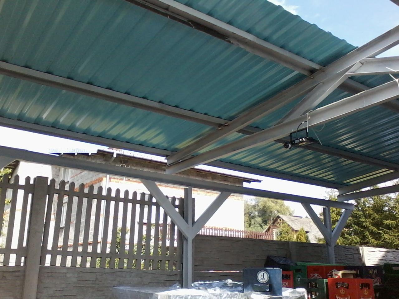 Zadaszenie z blachy falistej w kolorze turkusowym, wsparte na stalowej konstrukcji, widok z dołu, w tle betonowe ogrodzenie panelowe i budynek.