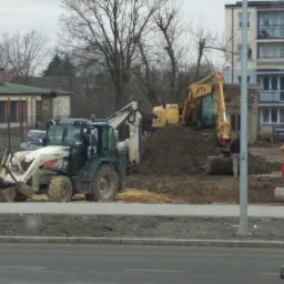 Prace ziemne w Łęcznej: koparko-ładowarka Terex i koparka Caterpillar pracują na placu budowy, widoczne hałdy ziemi i dwaj pracownicy w oddali.