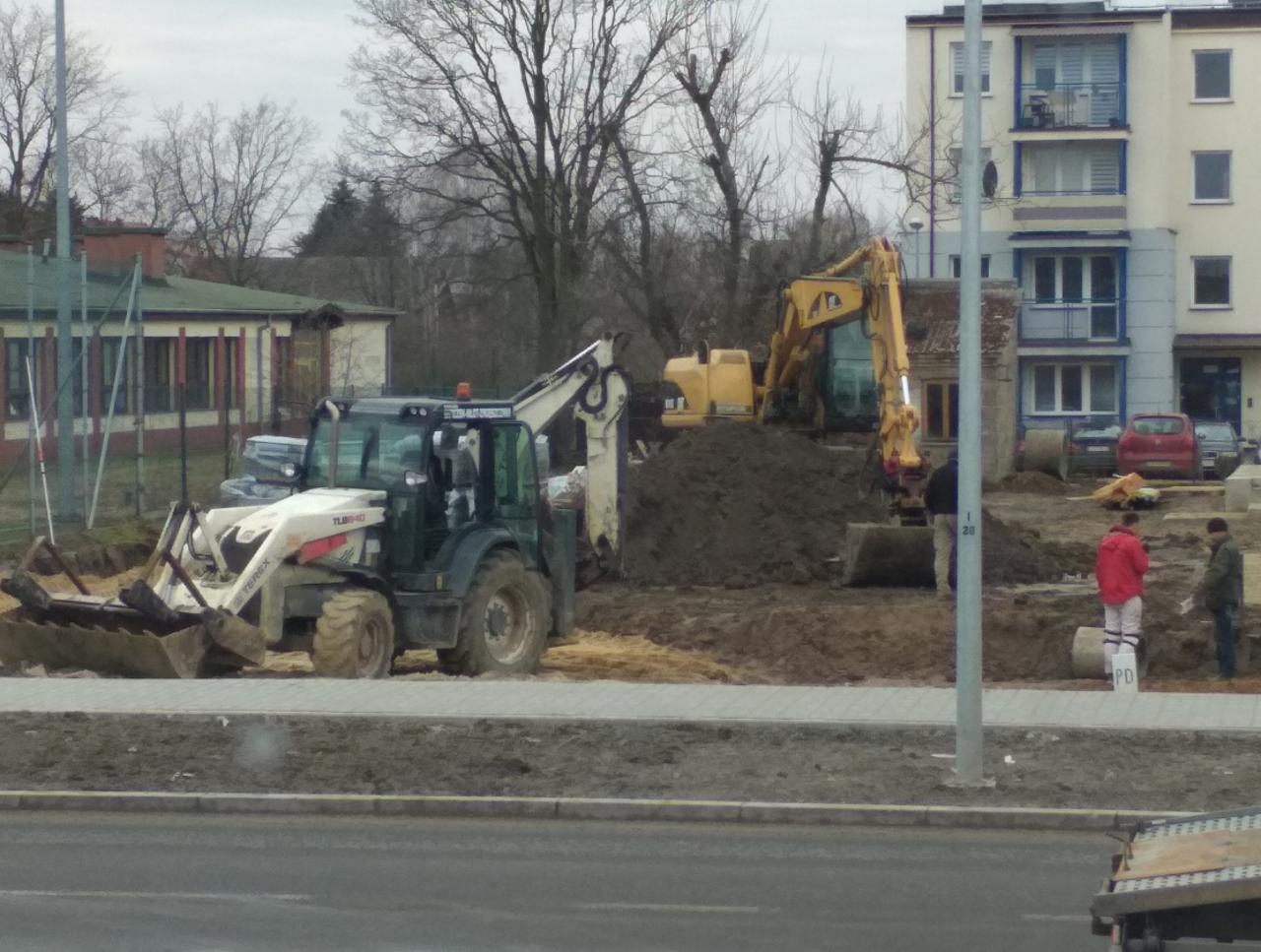 Prace ziemne w Łęcznej: koparko-ładowarka Terex i koparka Caterpillar pracują na placu budowy, widoczne hałdy ziemi i dwaj pracownicy w oddali.