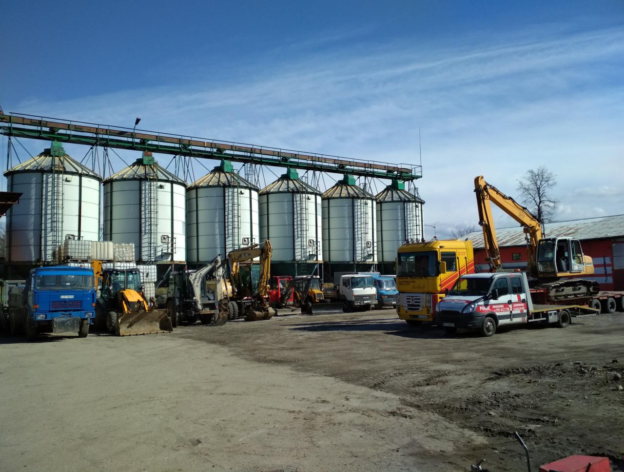 Różnorodny sprzęt budowlany, w tym koparki i ciężarówki, zaparkowany na placu budowy z rzędem silosów w tle pod błękitnym niebem.