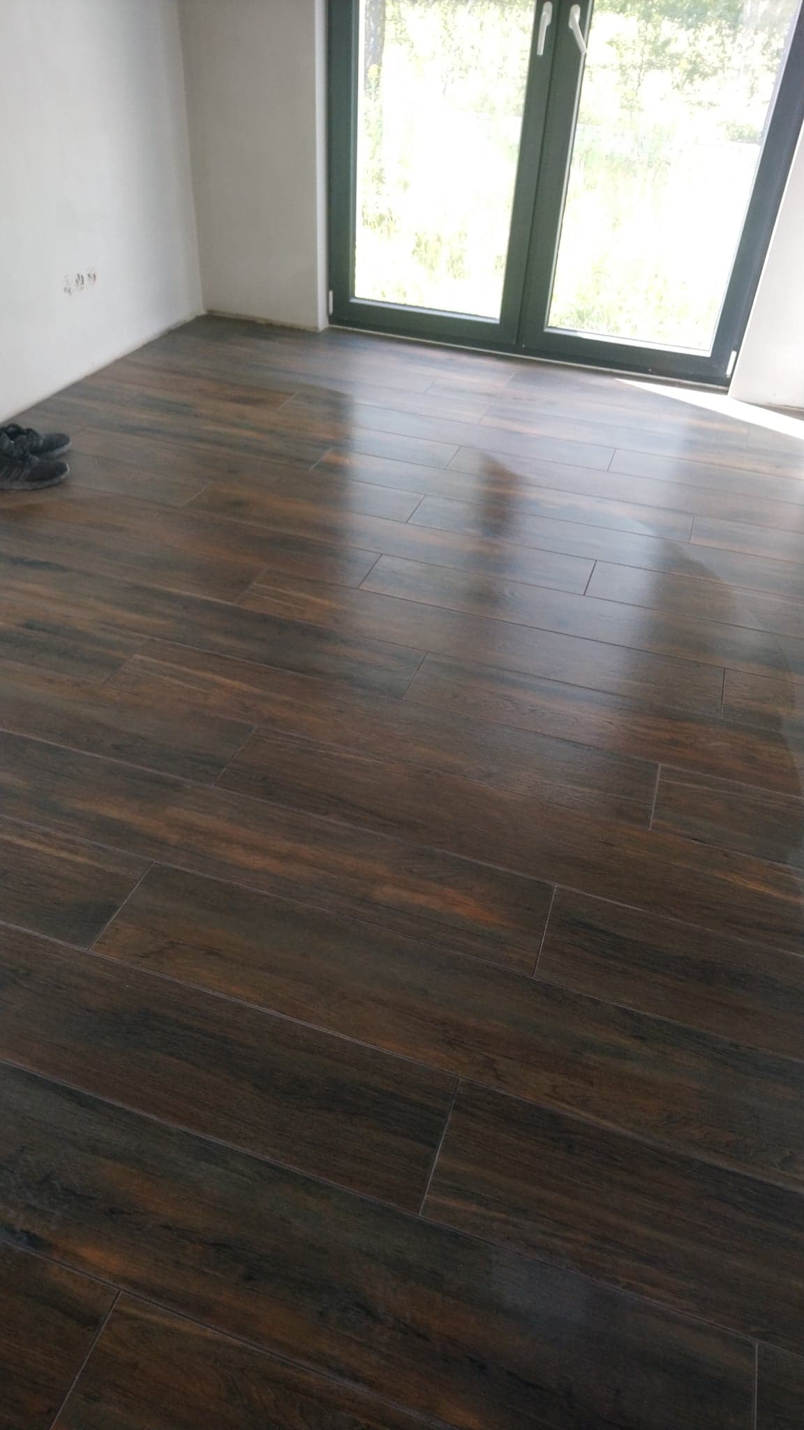 Ułożona, ciemnobrązowa, podłużna glazura imitująca drewno na podłodze w pustym pokoju z oknem i parą butów w rogu.
