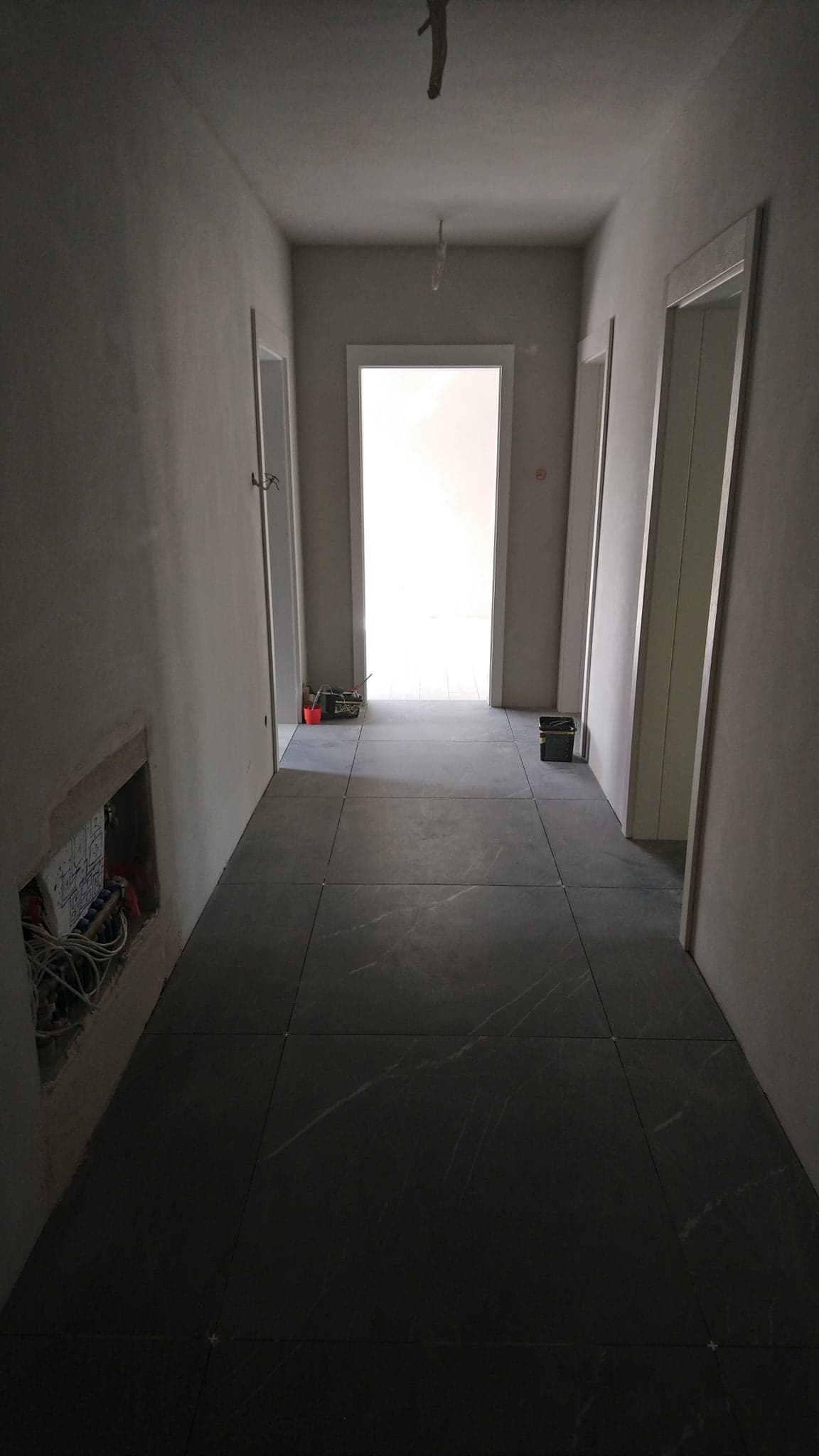 Korytarz w trakcie wykańczania, z szarymi płytkami podłogowymi imitującymi marmur, świeżo położone, ściany przygotowane pod wykończenie, widoczne puszki elektryczne i przewody, drzwi z białymi...