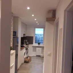 Widok na wyremontowaną kuchnię z perspektywy korytarza, z widoczną pralką, grzejnikiem pod oknem i rzędem punktowych lamp sufitowych.