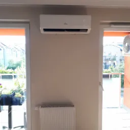 Biały klimatyzator marki Ferroli zamontowany na ścianie nad grzejnikiem, widok z wnętrza mieszkania na balkon z roślinami i osiedle.