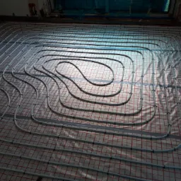 Układanie rur ogrzewania podłogowego na izolacji z siatką w pomieszczeniu w trakcie remontu, widoczne czarne klipsy mocujące rury.