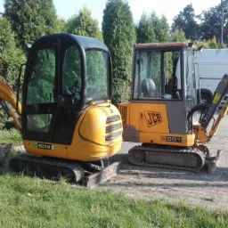 Dwie żółte mini koparki JCB, modele 8014 i 801, stoją obok siebie na trawie i kostce brukowej, z białym busem w tle.