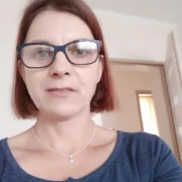 Portret kobiety z krótkimi, rudymi włosami i okularami w ciemnych oprawkach, noszącej srebrny łańcuszek z wisiorkiem, na neutralnym tle.