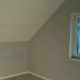 Pomalowane na szaro poddasze ze skośnym sufitem, widok z okna na budynek z wieżyczką w oddali.