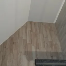 Wykończone poddasze z drewnopodobnymi panelami ściennymi i pomalowanym na biało skośnym sufitem, widoczny fragment drewnianej konstrukcji szkieletowej oraz czarny element instalacji.