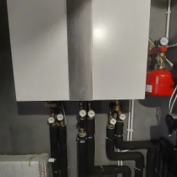 Nowoczesny, dwuczęściowy piec z logo Kostrzewa, z licznymi rurami i manometrami, zamontowany na szarej ścianie w kotłowni.