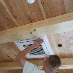 Monteraż sufitowej jednostki klimatyzacyjnej LG w pomieszczeniu z drewnianym sufitem i belkami; widoczna żarówka i otwór w suficie.