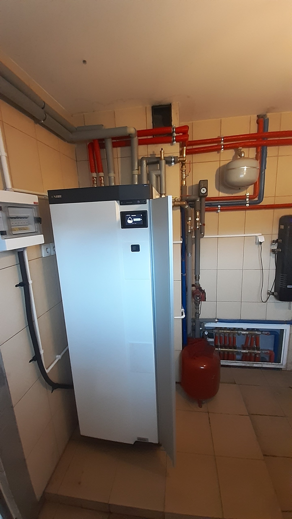 Biała pompa ciepła Nibe zamontowana w kotłowni, widoczne rury instalacji grzewczej i rozdzielacz podłogówki.