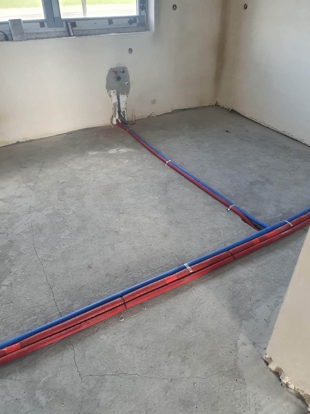 Nowa instalacja wodna w surowym wnętrzu: rury czerwone i niebieskie prowadzone po betonowej podłodze, mocowane metalowymi uchwytami, z wyprowadzeniami na ścianie pod oknem.