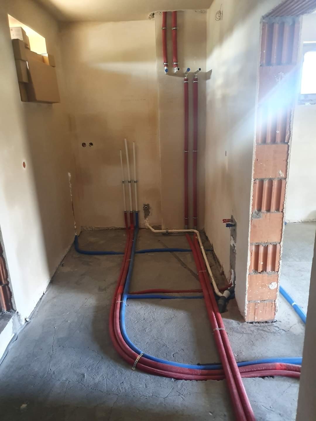 Surowy stan łazienki w trakcie instalacji hydraulicznej i grzewczej z widocznymi rurami PEX w kolorze niebieskim i czerwonym, prowadzonymi po podłodze i ścianach, oraz niezabezpieczonym murem...