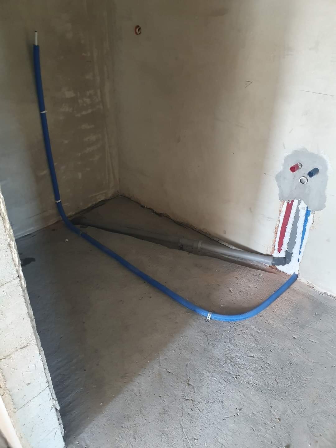 Niebieska rura instalacyjna przytwierdzona klamrami do podłogi w surowym pomieszczeniu, z widocznymi wyprowadzeniami rur w ścianie i szarym przewodem w tle.