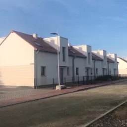 Szereg identycznych, białych domów jednorodzinnych z czerwonymi dachami, widocznych od strony ulicy z chodnikiem z kostki brukowej i przygotowywanym podłożem pod drogę.