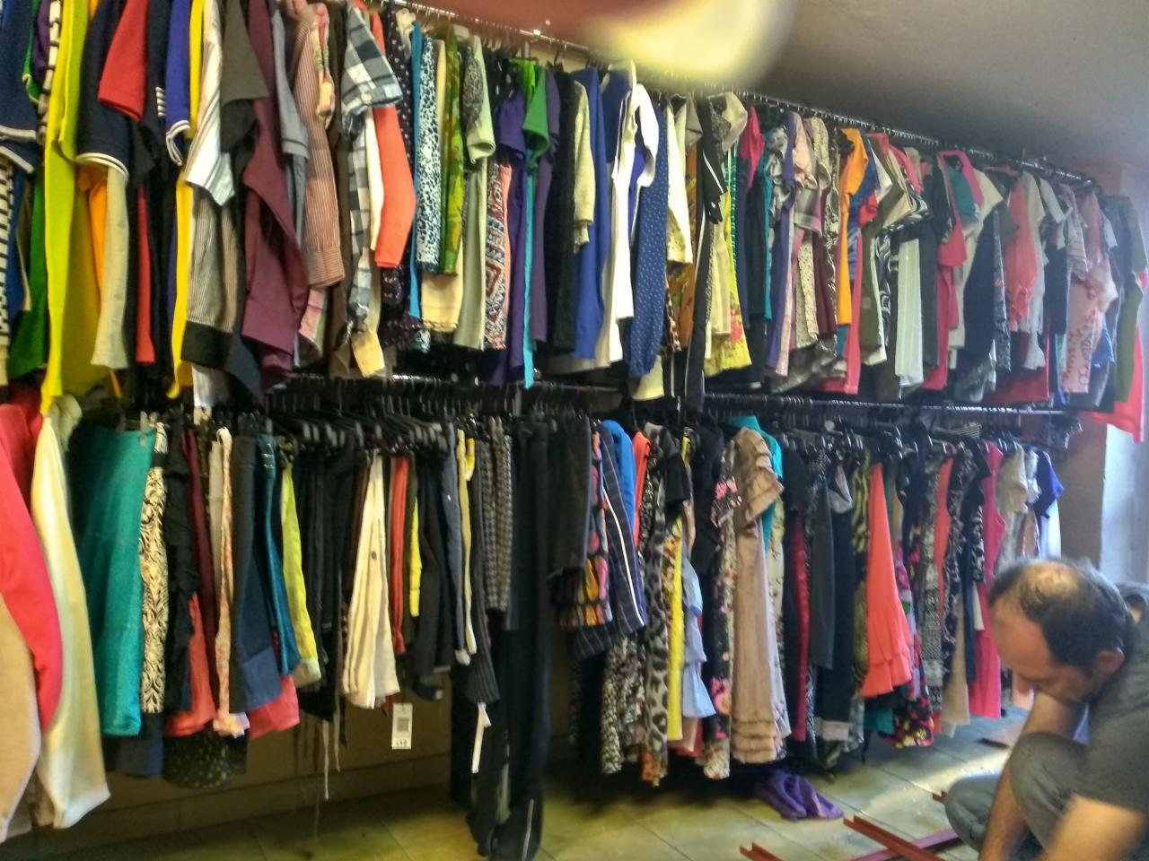 Rzędy ubrań wiszące na wieszakach w sklepie, widoczny mężczyzna siedzący na podłodze, prawdopodobnie pracownik.