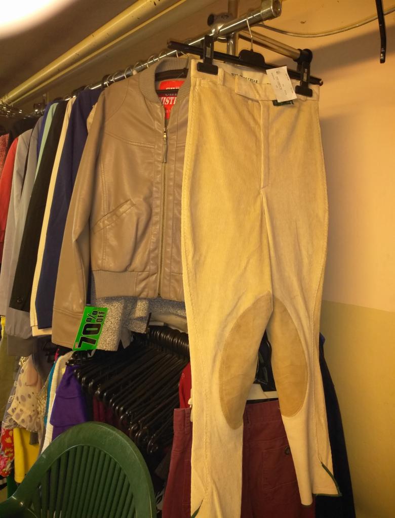 Beżowe spodnie bryczesy z łatami na kolanach i zielonym wykończeniem u dołu, wiszące na wieszaku obok beżowej kurtki i innych ubrań na metalowym drążku w sklepie z odzieżą używaną, widoczna...