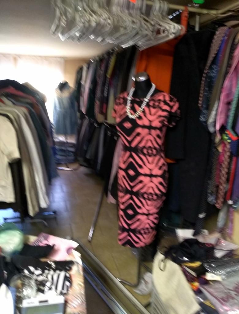 Wnętrze sklepu z odzieżą, z wieszakami u góry i manekinem w sukience z geometrycznym wzorem w kolorze koralowym i czarnym, uzupełnionym o sznur pereł.