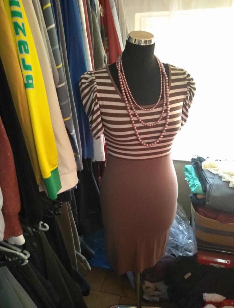 Sukienka w paski na manekinie z trzema sznurami różowych pereł, otoczona ubraniami na wieszakach i w pudłach.