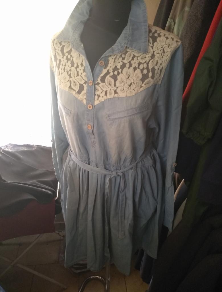 Jasnoniebieska sukienka koszulowa z koronkową wstawką na gorsecie, zawiązywana w pasie, prezentowana na manekinie na tle ubrań.