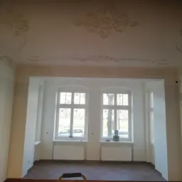 Wnętrze odnowionego pokoju z parkietem w jodełkę, białymi ścianami z dekoracyjnymi sztukateriami na suficie, dwoma oknami z widokiem na zewnątrz i drabinką malarską na pierwszym planie.