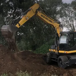 Żółta koparka New Holland WE150B PRO wysypuje ziemię z łyżki na pryzmę ziemi w trakcie prac ziemnych, w tle drzewa.