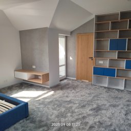 MAX MAREK DOMINIK KANDZIA - Jasne wnętrze pokoju na poddaszu z szarą, teksturowaną ścianą, drewnianymi elementami i niebieskim łóżkiem bez materaca, ukazujące etap wykończenia przed umeblowaniem.