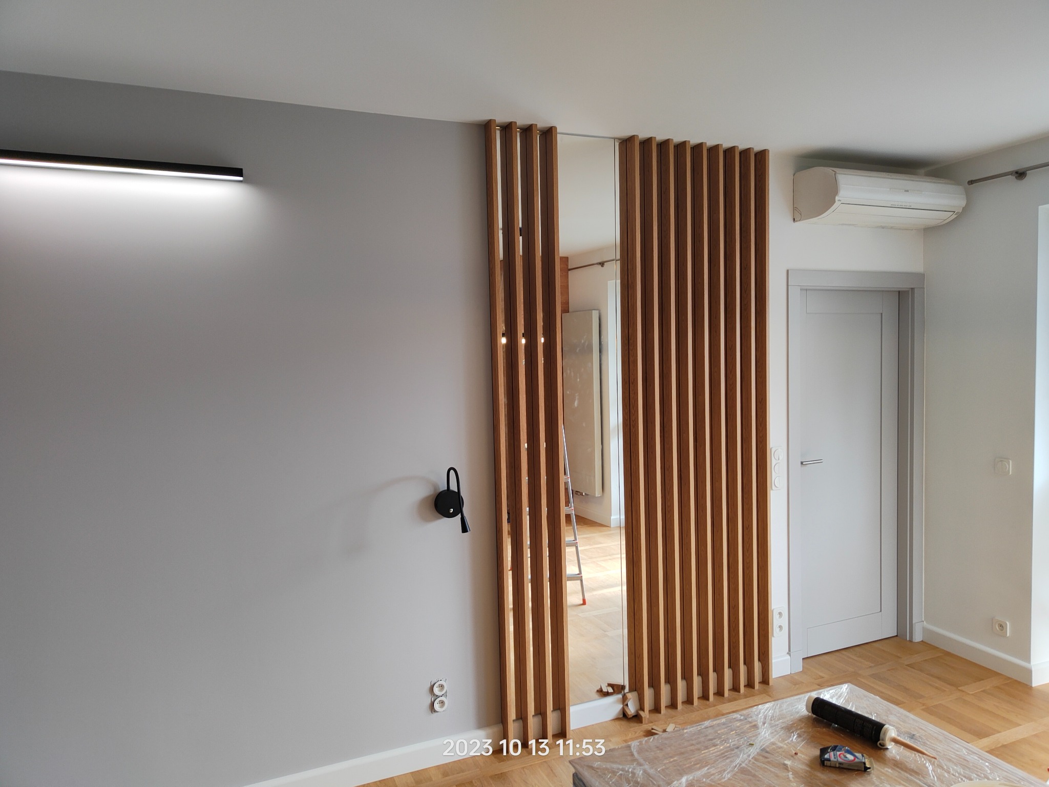 Montaż drewnianych lameli dekoracyjnych w przedpokoju, widoczna lampa ścienna, gniazdka elektryczne i częściowo zakryte meble folią ochronną.