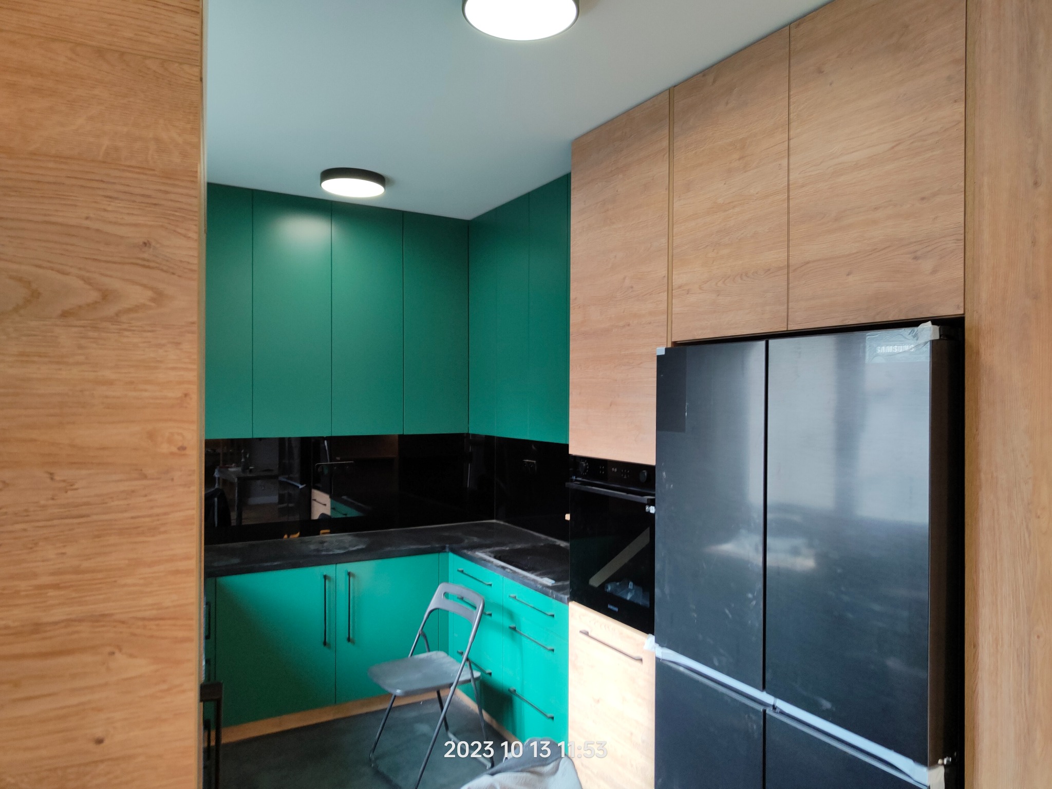 Nowoczesna kuchnia z zielonymi szafkami dolnymi i drewnopodobnymi górnymi, czarnym blatem i lodówką Samsung, widok na krzesło składane.