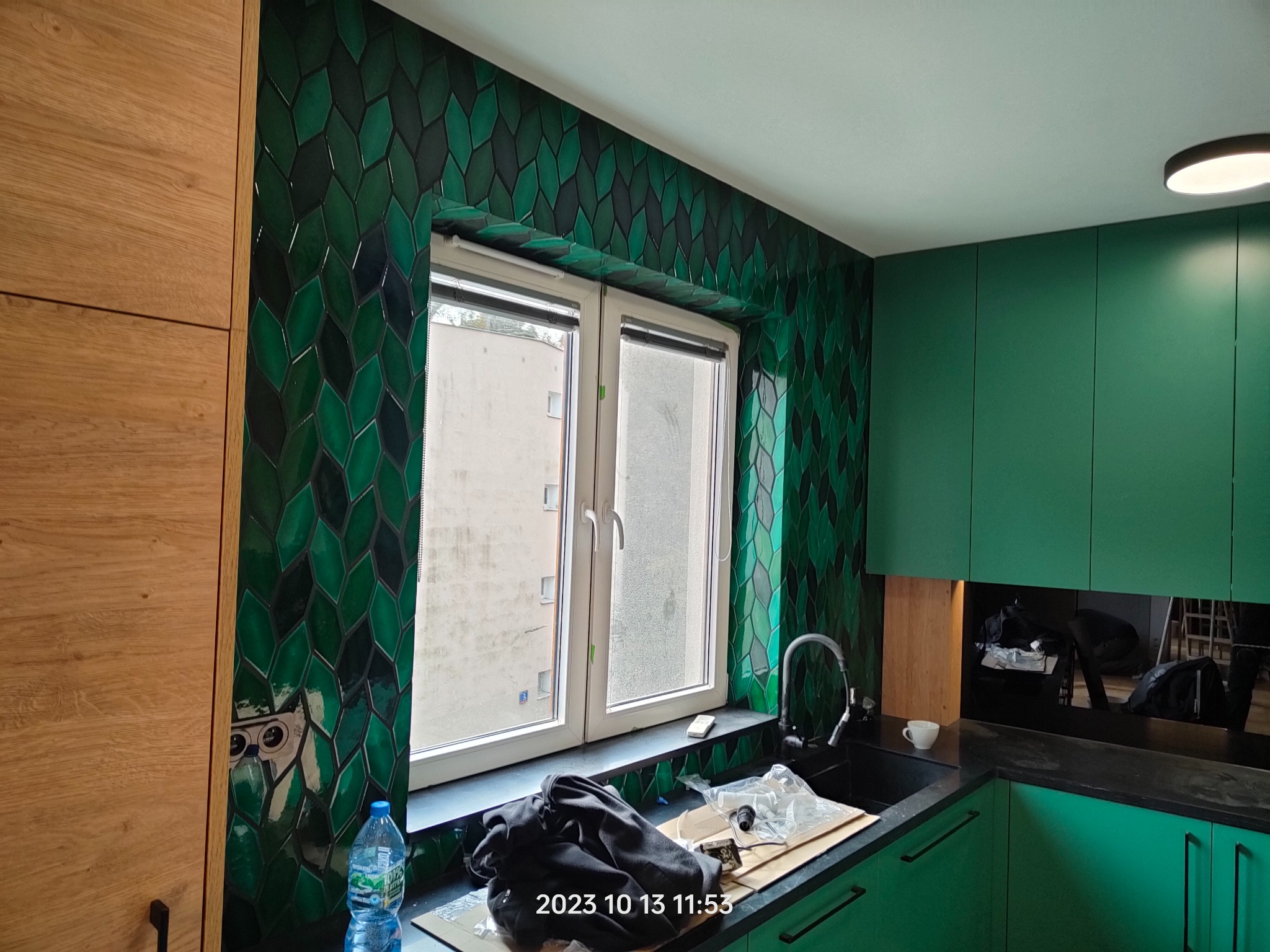 Kuchnia w trakcie wykańczania: zielone, łezkowate płytki na ścianie, okno z widokiem na budynek, czarny zlew i zielone szafki. Widać butelkę wody i ubrania na blacie.