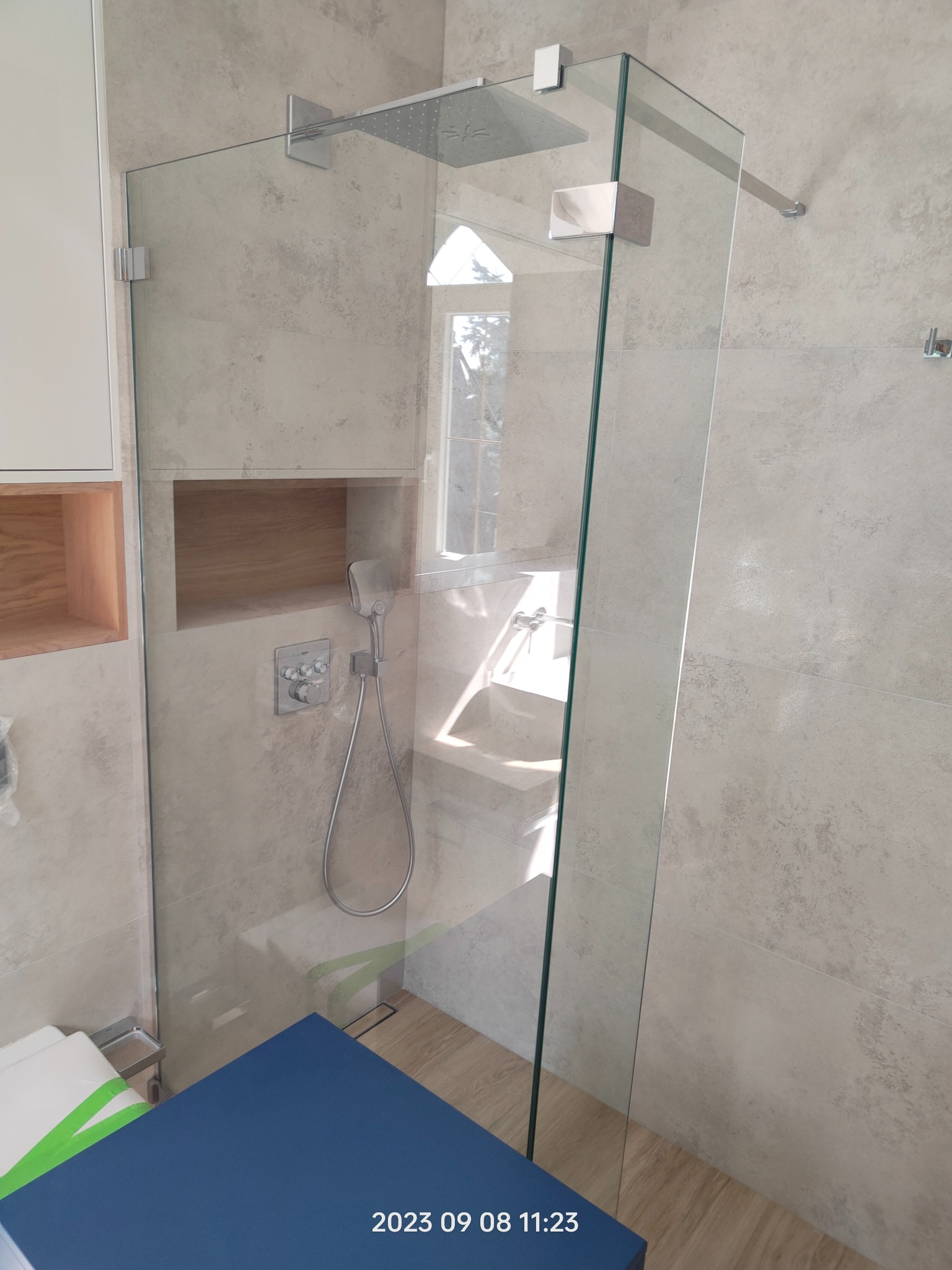 Nowoczesna kabina prysznicowa walk-in z transparentnego szkła, zamocowana na chromowanych uchwytach, z deszczownicą i panelem prysznicowym na ścianie z beżowych płytek imitujących kamień, w tle...