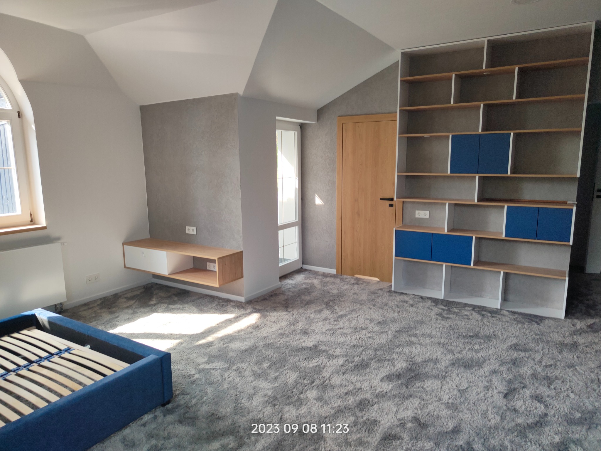 Jasne wnętrze pokoju na poddaszu z szarą, teksturowaną ścianą, drewnianymi elementami i niebieskim łóżkiem bez materaca, ukazujące etap wykończenia przed umeblowaniem.